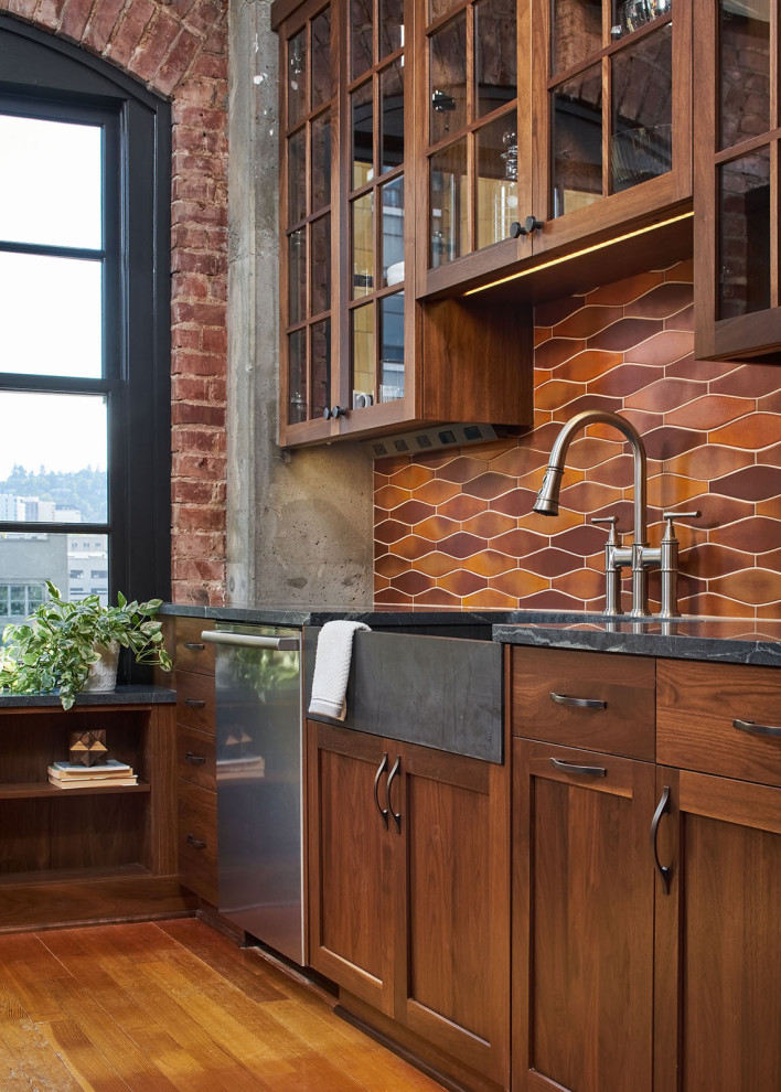 This is an example of a mid-sized industrial l-shaped open plan kitchen in Portland with a farmhouse sink, glass-front cabinets, dark wood cabinets, soapstone benchtops, multi-coloured splashback, ceramic splashback, stainless steel appliances, medium hardwood floors, with island, brown floor, black benchtop and exposed beam.