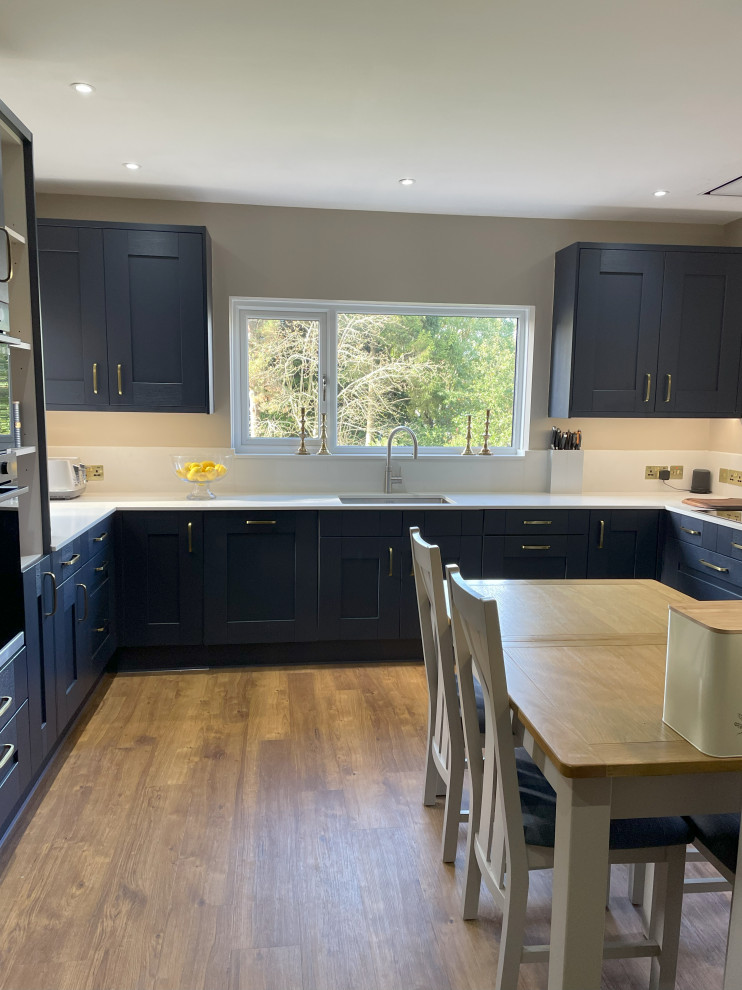 Medium sized classic u-shaped kitchen/diner in Other with an integrated sink, shaker cabinets, blue cabinets, quartz worktops, white splashback, black appliances, laminate floors, no island, brown floors, white worktops and feature lighting.