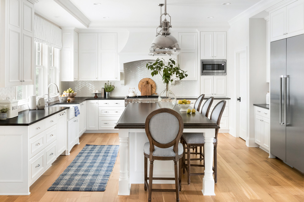 This is an example of a large beach style l-shaped eat-in kitchen in Minneapolis with a farmhouse sink, white cabinets, quartz benchtops, white splashback, mosaic tile splashback, stainless steel appliances, with island, brown floor, recessed-panel cabinets, medium hardwood floors and black benchtop.