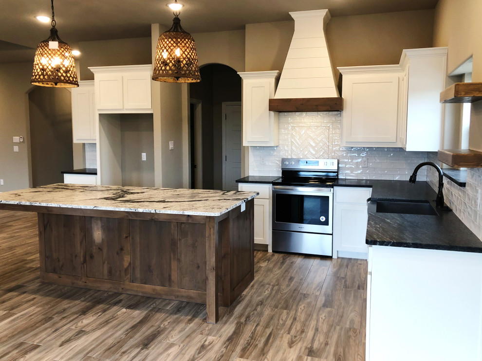 Mid-sized country u-shaped open plan kitchen in Dallas with a farmhouse sink, shaker cabinets, white cabinets, granite benchtops, white splashback, subway tile splashback, stainless steel appliances, with island, brown floor and black benchtop.
