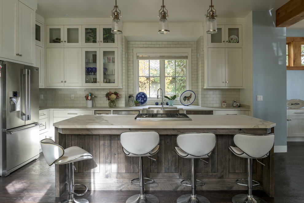 Inspiration for a country l-shaped kitchen in Boise with a farmhouse sink, shaker cabinets, white cabinets, white splashback, subway tile splashback, stainless steel appliances, dark hardwood floors, with island, brown floor and white benchtop.