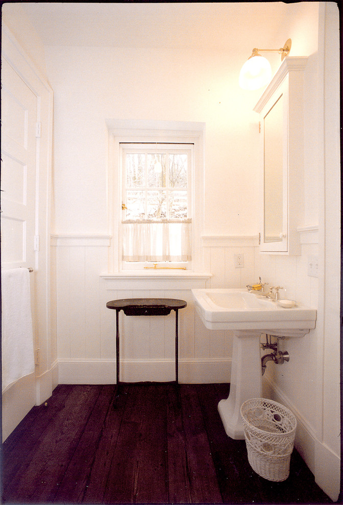 This is an example of a mid-sized country 3/4 bathroom in New York with a pedestal sink, recessed-panel cabinets, white cabinets, an undermount tub, a shower/bathtub combo, a two-piece toilet, white walls and dark hardwood floors.