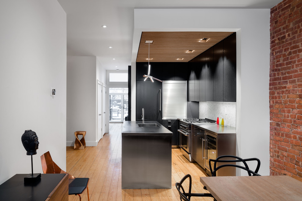 Small midcentury l-shaped eat-in kitchen in New York with an integrated sink, flat-panel cabinets, black cabinets, stainless steel benchtops, white splashback, stone tile splashback, stainless steel appliances, light hardwood floors and with island.