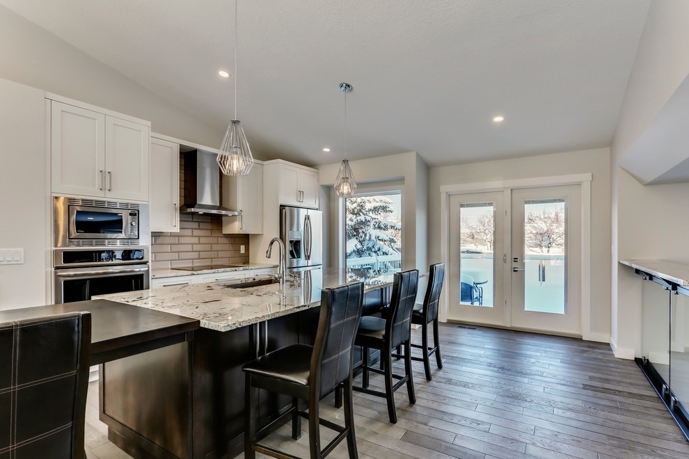 Inspiration for a large modern eat-in kitchen in Calgary with a drop-in sink, white cabinets, granite benchtops, grey splashback, subway tile splashback, stainless steel appliances, medium hardwood floors, with island and brown floor.