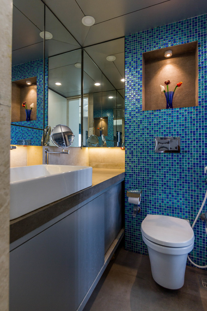 This is an example of a contemporary bathroom in Mumbai with flat-panel cabinets, grey cabinets, a wall-mount toilet, blue tile, green tile, glass tile, a vessel sink, grey floor and grey benchtops.