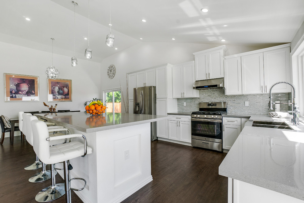 This is an example of a large modern l-shaped eat-in kitchen in Seattle with a farmhouse sink, shaker cabinets, white cabinets, quartz benchtops, grey splashback, glass sheet splashback, stainless steel appliances, laminate floors, with island, brown floor and grey benchtop.