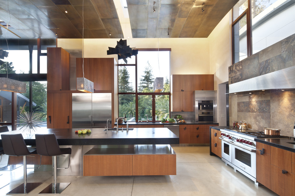 This is an example of a large contemporary l-shaped open plan kitchen in San Francisco with stainless steel appliances, flat-panel cabinets, medium wood cabinets, soapstone benchtops, brown splashback, an undermount sink, slate splashback, concrete floors and with island.