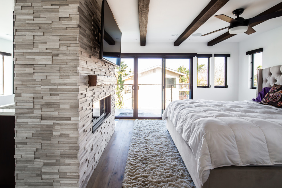 Large beach style master bedroom in San Diego with white walls, laminate floors, a two-sided fireplace, a tile fireplace surround and brown floor.
