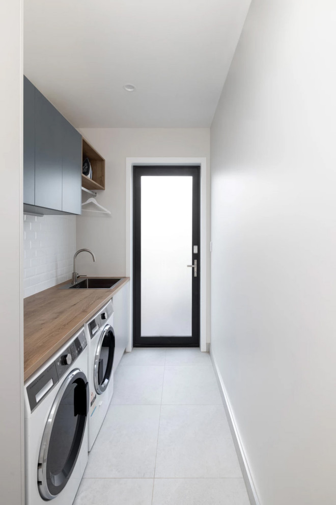 Photo of a mid-sized contemporary single-wall dedicated laundry room in Auckland with white splashback, ceramic splashback, white benchtop, a drop-in sink, flat-panel cabinets, blue cabinets, wood benchtops, white walls, porcelain floors and a side-by-side washer and dryer.