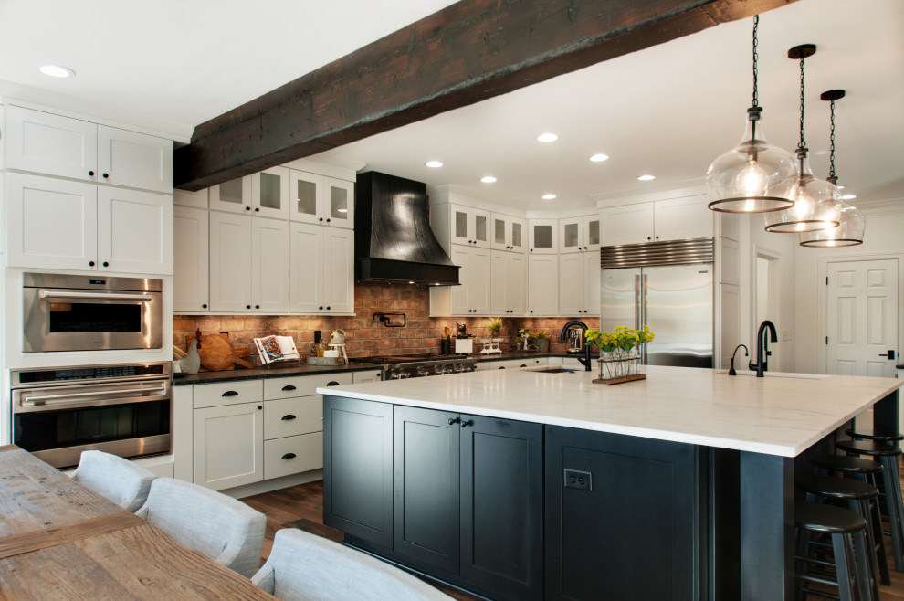 Photo of a large country u-shaped open plan kitchen in Nashville with an undermount sink, shaker cabinets, white cabinets, quartz benchtops, multi-coloured splashback, brick splashback, stainless steel appliances, medium hardwood floors, with island, brown floor, white benchtop and exposed beam.