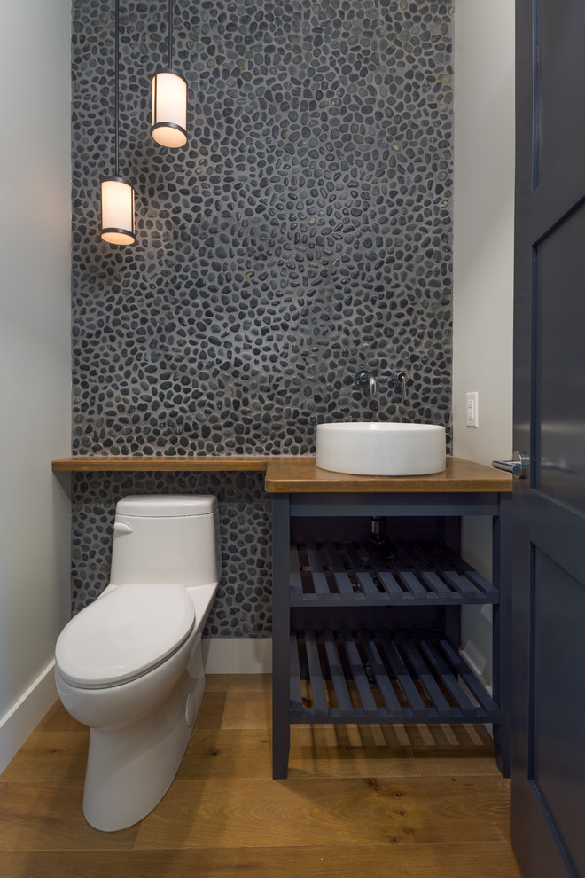 Photo of a mid-sized modern powder room with a one-piece toilet, gray tile, pebble tile, white walls, medium hardwood floors, a vessel sink, brown floor and brown benchtops.