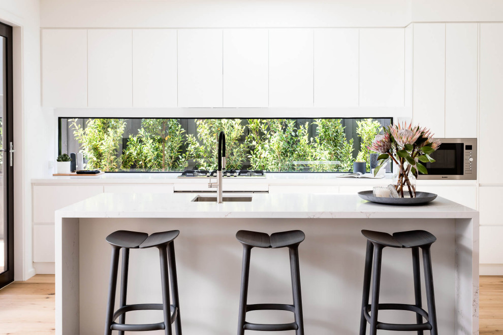 Photo of a contemporary galley kitchen in Melbourne with an undermount sink, flat-panel cabinets, white cabinets, window splashback, medium hardwood floors, with island, brown floor and white benchtop.