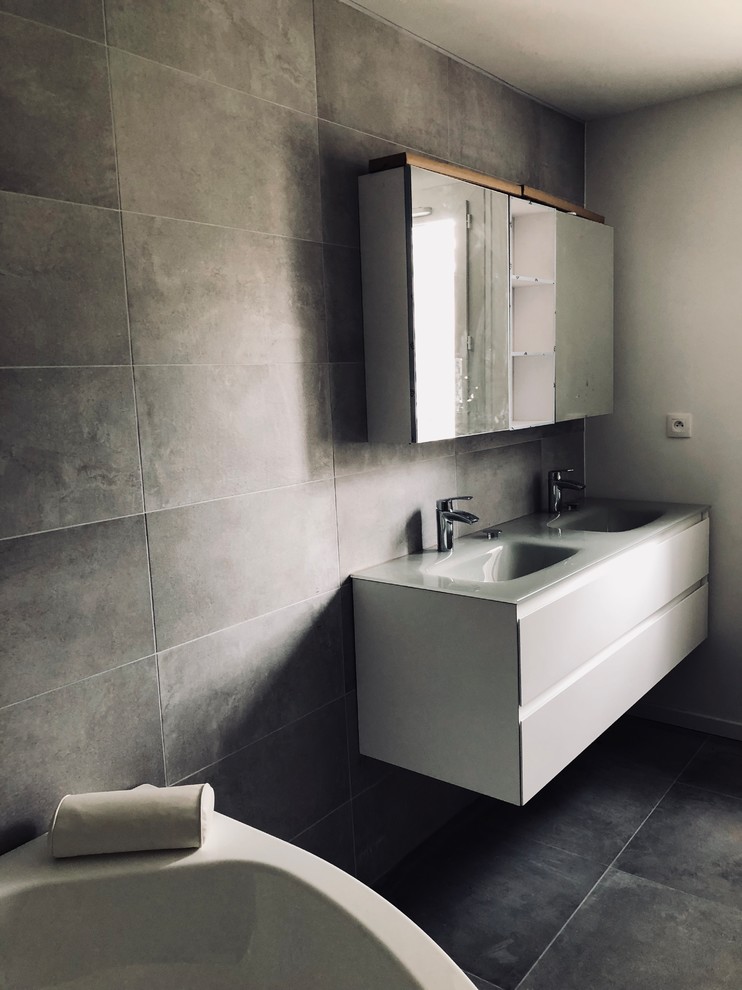Large contemporary master bathroom in Paris with a hot tub, a curbless shower, ceramic tile, ceramic floors, grey floor, an open shower, flat-panel cabinets, white cabinets, a two-piece toilet, gray tile, grey walls, a wall-mount sink, glass benchtops and grey benchtops.