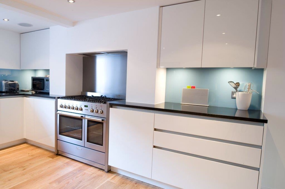 Large contemporary u-shaped eat-in kitchen in London with flat-panel cabinets, white cabinets, granite benchtops, blue splashback, glass sheet splashback, stainless steel appliances, light hardwood floors and no island.