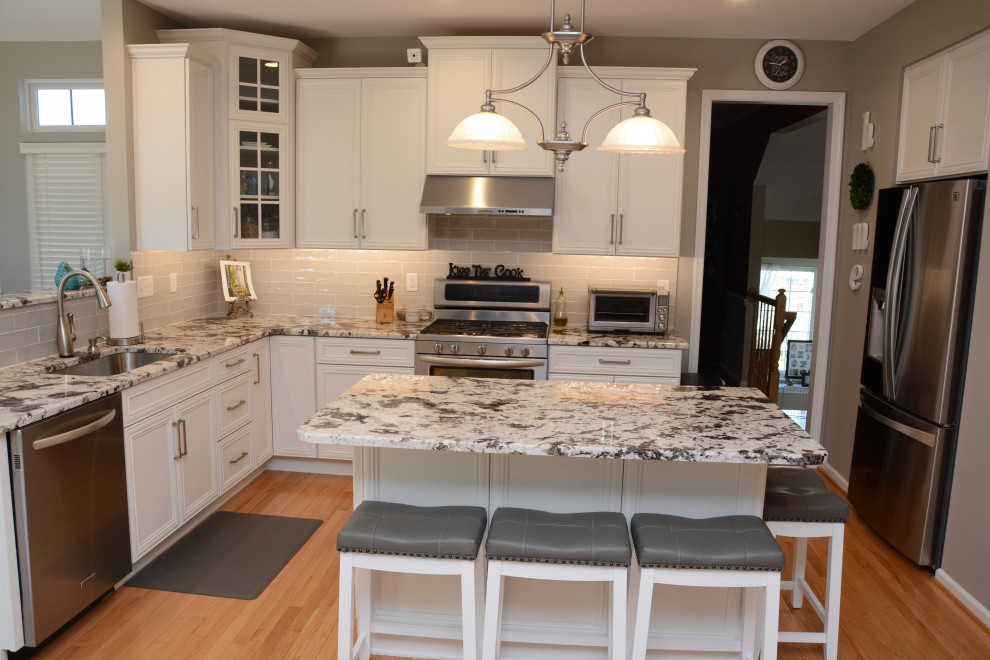 Inspiration for a mid-sized traditional u-shaped open plan kitchen in DC Metro with an undermount sink, recessed-panel cabinets, white cabinets, granite benchtops, grey splashback, stainless steel appliances, light hardwood floors, with island, brown floor and white benchtop.
