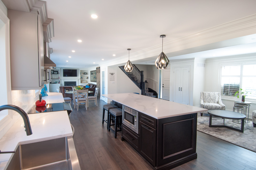 This is an example of a large contemporary l-shaped eat-in kitchen in Toronto with a farmhouse sink, flat-panel cabinets, grey cabinets, quartz benchtops, white splashback, ceramic splashback, stainless steel appliances, medium hardwood floors, with island, brown floor and white benchtop.