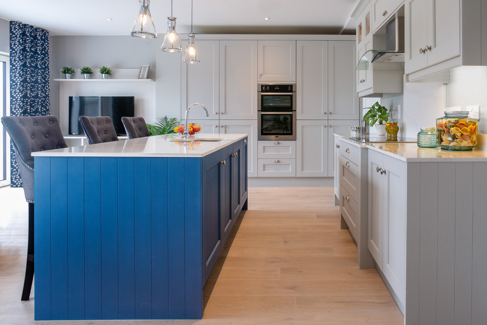 This is an example of a large transitional l-shaped kitchen in Dublin with recessed-panel cabinets, grey cabinets, quartzite benchtops, white splashback, panelled appliances, light hardwood floors, with island, white benchtop, an undermount sink, stone slab splashback and beige floor.