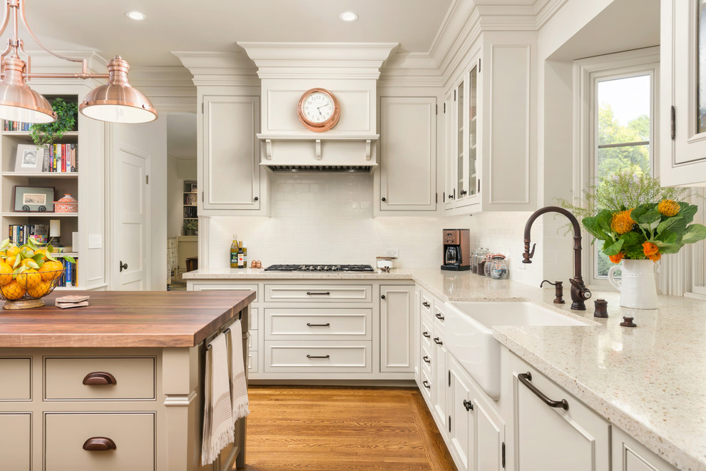 Photo of a mid-sized mediterranean l-shaped open plan kitchen in San Francisco with a farmhouse sink, recessed-panel cabinets, white cabinets, terrazzo benchtops, white splashback, subway tile splashback, panelled appliances, medium hardwood floors, with island, brown floor and beige benchtop.