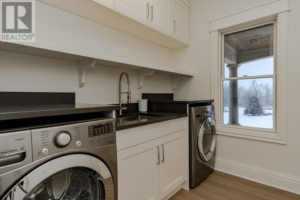 Design ideas for a mid-sized transitional single-wall dedicated laundry room in Ottawa with an undermount sink, shaker cabinets, white cabinets, quartz benchtops, white walls, light hardwood floors, an integrated washer and dryer and black benchtop.