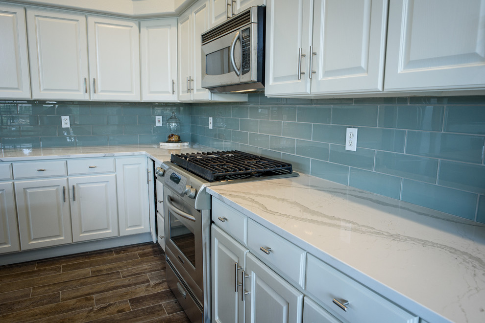 This is an example of a large industrial l-shaped open plan kitchen in Denver with a farmhouse sink, raised-panel cabinets, white cabinets, blue splashback, glass tile splashback, stainless steel appliances, porcelain floors, with island and quartz benchtops.