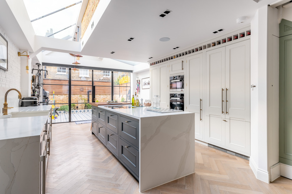 Photo of a large traditional galley open plan kitchen in London with a built-in sink, recessed-panel cabinets, beige cabinets, marble worktops, white splashback, marble splashback, black appliances, light hardwood flooring, an island, brown floors, white worktops, a vaulted ceiling and feature lighting.