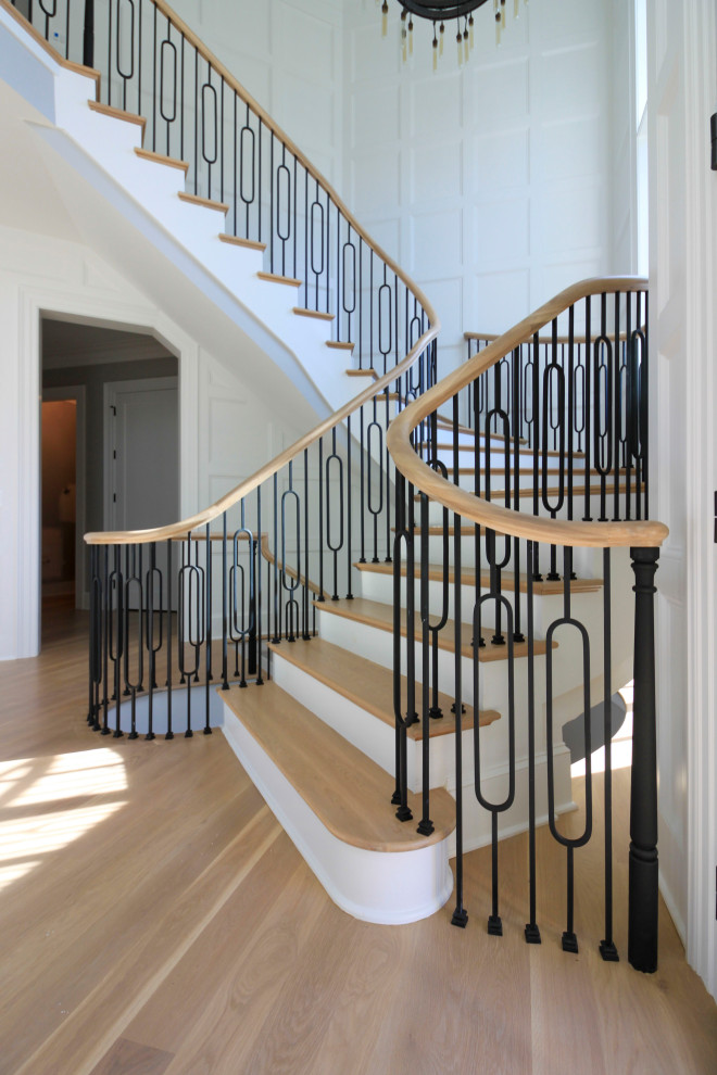 Пример оригинального дизайна: огромная изогнутая деревянная лестница в стиле неоклассика (современная классика) с деревянными ступенями, перилами из смешанных материалов и панелями на стенах