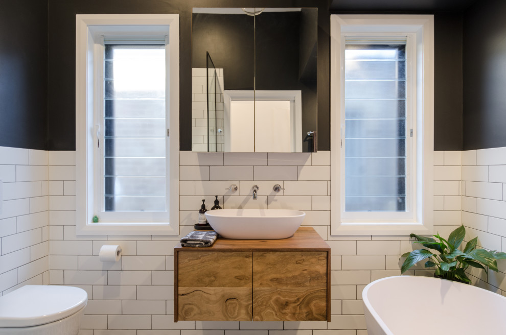 Photo of a contemporary 3/4 bathroom in Sydney with flat-panel cabinets, brown cabinets, a freestanding tub, an alcove shower, a wall-mount toilet, white tile, black walls, a vessel sink, wood benchtops, brown benchtops and a single vanity.