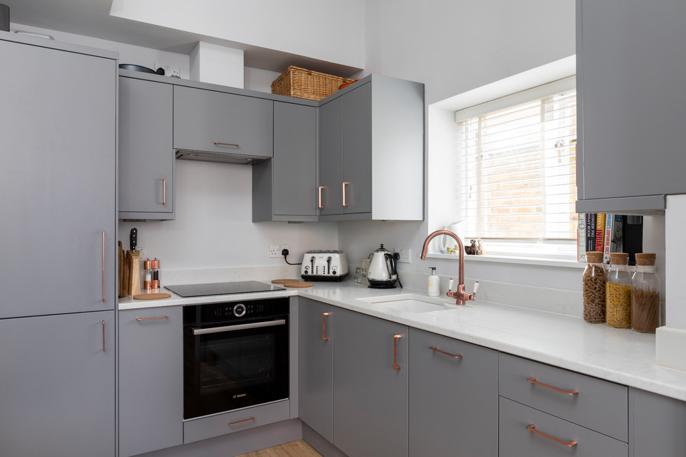 This is an example of a small arts and crafts l-shaped open plan kitchen in London with an integrated sink, flat-panel cabinets, grey cabinets, solid surface benchtops, white splashback, limestone splashback, stainless steel appliances, laminate floors and no island.