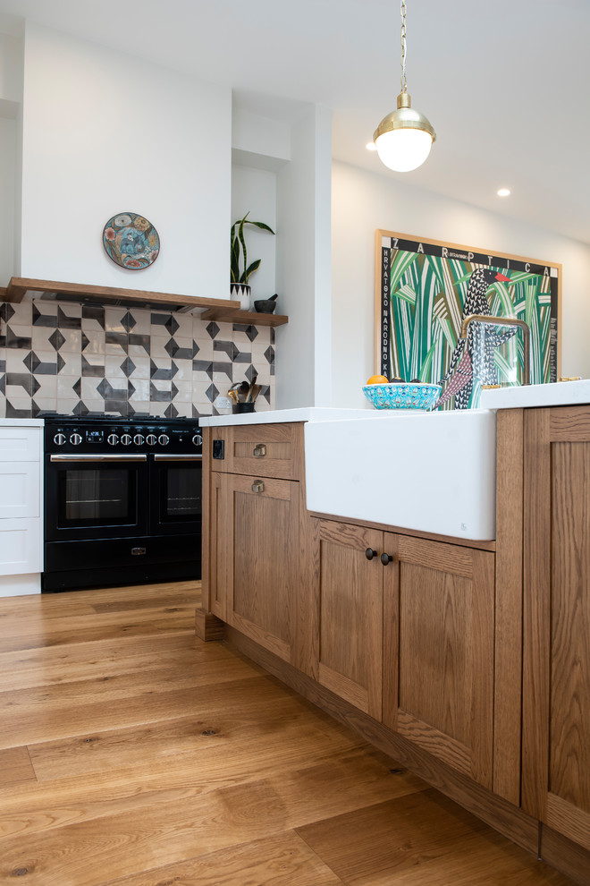 Photo of a large mediterranean l-shaped open plan kitchen in Melbourne with a farmhouse sink, shaker cabinets, quartz benchtops, grey splashback, cement tile splashback, black appliances, medium hardwood floors, with island, white benchtop and medium wood cabinets.