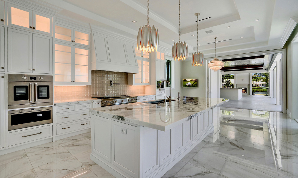This is an example of an expansive mediterranean kitchen in Miami with an undermount sink, white cabinets, marble benchtops, beige splashback, ceramic splashback, stainless steel appliances, with island, white floor, white benchtop and glass-front cabinets.