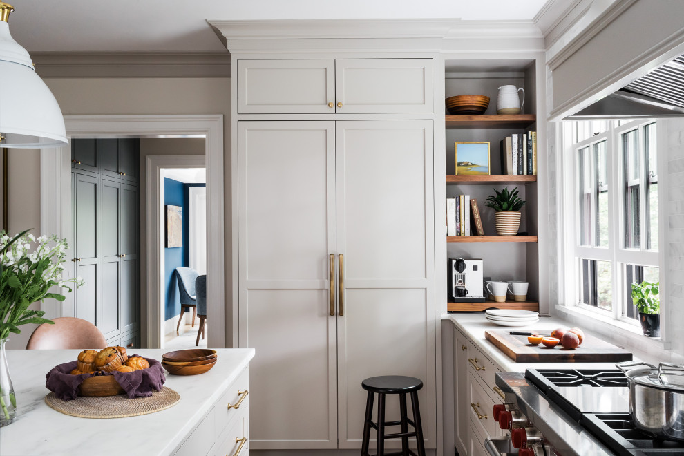 Inredning av ett klassiskt vit vitt kök, med en rustik diskho, luckor med glaspanel, beige skåp, marmorbänkskiva, vitt stänkskydd, stänkskydd i marmor, rostfria vitvaror, ljust trägolv och en köksö