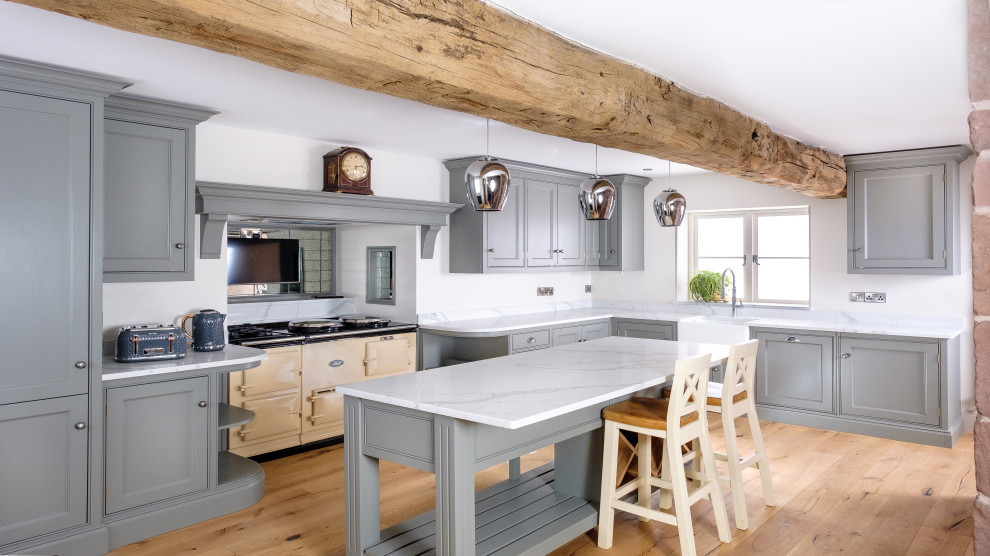 This is an example of a farmhouse l-shaped kitchen with a belfast sink, beaded cabinets, grey cabinets, coloured appliances, light hardwood flooring, an island, beige floors, white worktops and exposed beams.