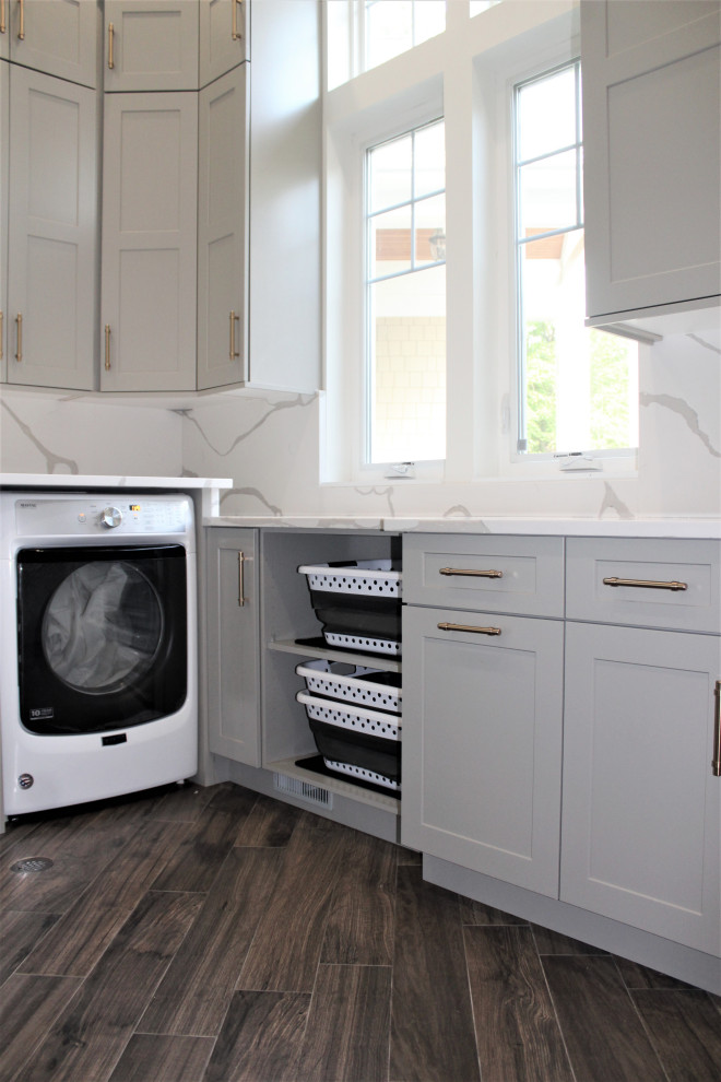 This is an example of a large classic u-shaped separated utility room in Detroit with a belfast sink, shaker cabinets, grey cabinets, engineered stone countertops, white splashback, engineered quartz splashback, grey walls, dark hardwood flooring, a side by side washer and dryer, brown floors and white worktops.