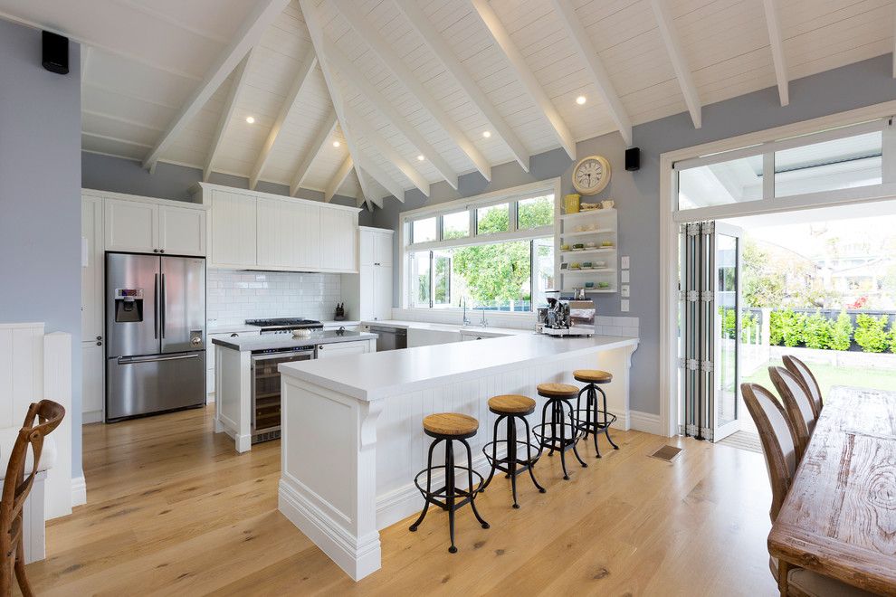 This is an example of a Mid-sized transitional U-shaped eat-in kitchen in Hamilton With Shaker Cabinets, White Cabinets, Quartz Benchtops, White Splashback, Ceramic Splashback, White Appliances, with Island, White Benchtop, a Farmhouse Sink, Medium Hardwood Floors and Brown Floor.