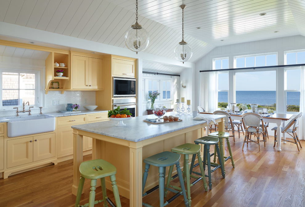 Foto på ett maritimt vit kök och matrum, med en rustik diskho, gula skåp, marmorbänkskiva, vitt stänkskydd, stänkskydd i stenkakel, integrerade vitvaror, ljust trägolv, en köksö och luckor med infälld panel