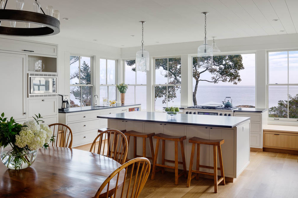 This is an example of a beach style l-shaped eat-in kitchen in Boston with shaker cabinets, white cabinets, panelled appliances, with island and light hardwood floors.