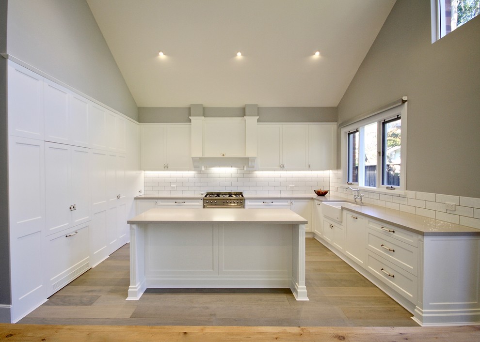 This is an example of a large transitional l-shaped eat-in kitchen in Sydney with a farmhouse sink, shaker cabinets, white cabinets, quartz benchtops, white splashback, ceramic splashback, stainless steel appliances, light hardwood floors, with island and brown floor.