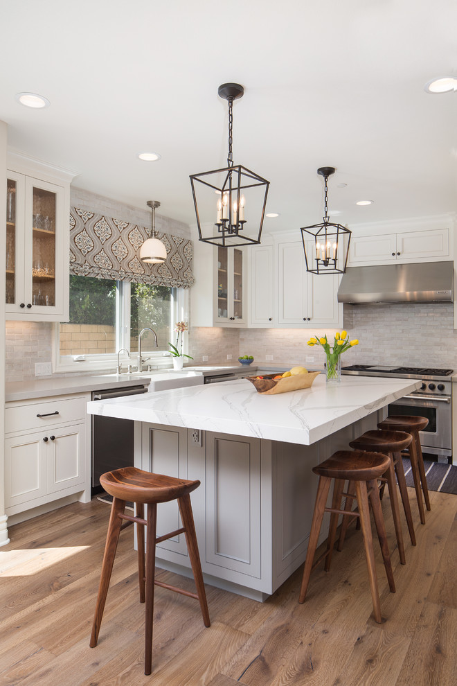 This is an example of a mid-sized transitional u-shaped separate kitchen in Los Angeles with a farmhouse sink, recessed-panel cabinets, white cabinets, marble benchtops, grey splashback, stainless steel appliances, dark hardwood floors, with island, brown floor, white benchtop and subway tile splashback.