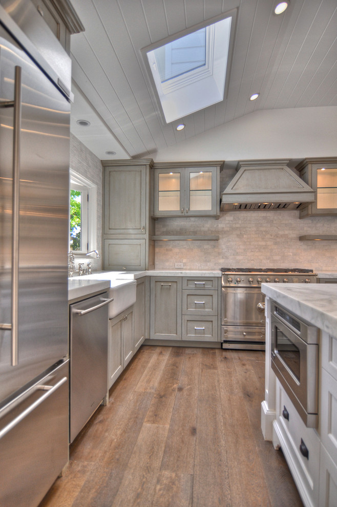 This is an example of a large country u-shaped eat-in kitchen in Orange County with a farmhouse sink, recessed-panel cabinets, distressed cabinets, marble benchtops, grey splashback, stainless steel appliances, stone tile splashback, medium hardwood floors and with island.