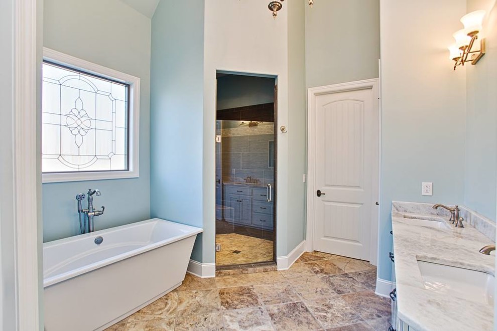 Mid-sized transitional master beige tile and ceramic tile travertine floor freestanding bathtub photo with raised-panel cabinets, distressed cabinets, blue walls, an undermount sink and marble countertops