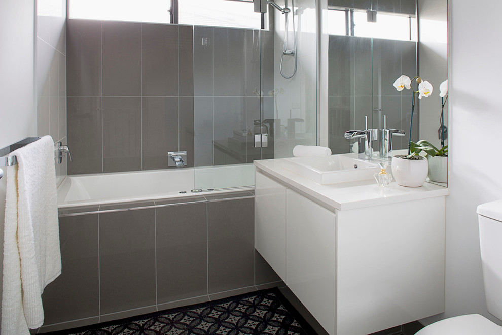 This is an example of a small contemporary 3/4 bathroom in Melbourne with flat-panel cabinets, white cabinets, a drop-in tub, a shower/bathtub combo, a one-piece toilet, gray tile, porcelain tile, white walls, ceramic floors, a wall-mount sink, grey floor and a hinged shower door.