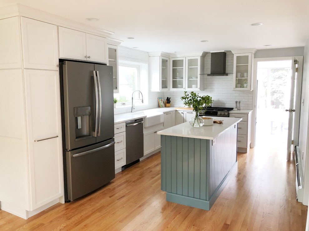 Mid-sized transitional l-shaped light wood floor kitchen photo in Providence with a farmhouse sink, shaker cabinets, white cabinets, quartz countertops, white backsplash, ceramic backsplash, stainless steel appliances, an island and white countertops
