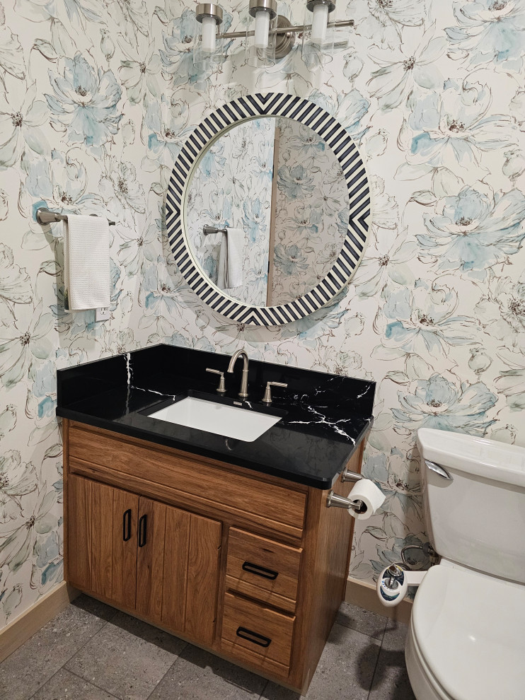 Photo of a mid-sized transitional powder room in Other with medium wood cabinets, porcelain floors, glass benchtops, grey floor, black benchtops, a built-in vanity and wallpaper.