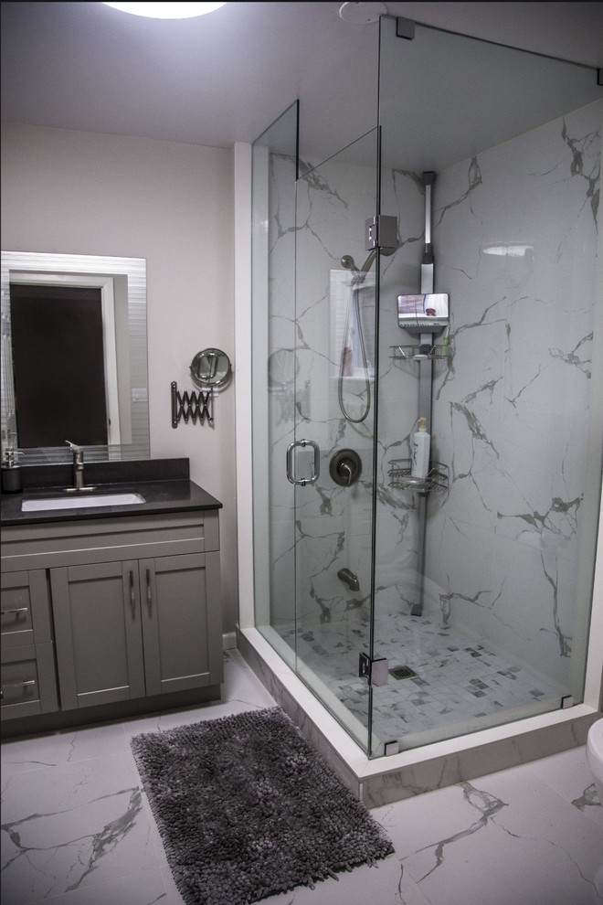 This is an example of a mid-sized transitional 3/4 bathroom in Toronto with shaker cabinets, grey cabinets, a corner shower, white tile, grey walls, an undermount sink, granite benchtops, white floor, a hinged shower door and black benchtops.