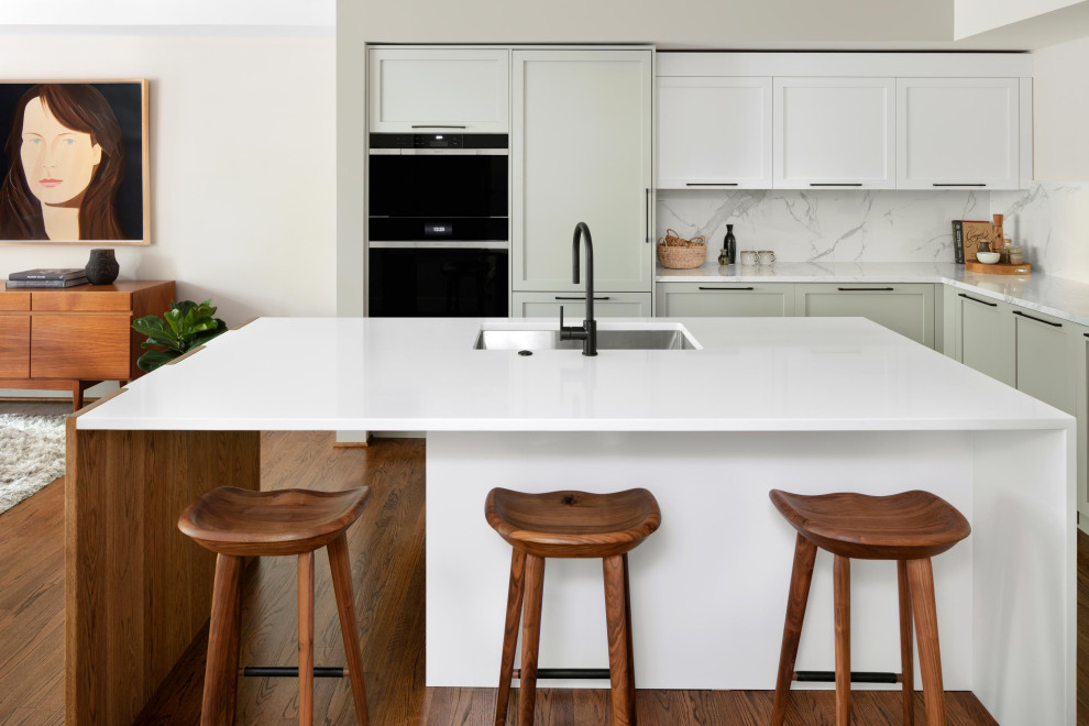 Example of a large trendy l-shaped medium tone wood floor and brown floor open concept kitchen design in DC Metro with an undermount sink, recessed-panel cabinets, gray cabinets, quartz countertops, white backsplash, porcelain backsplash, black appliances, an island and white countertops