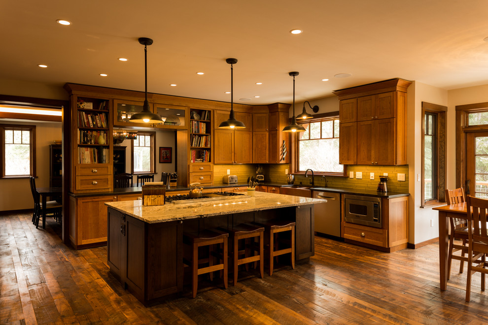 Example of a large mountain style u-shaped dark wood floor and brown floor eat-in kitchen design with a farmhouse sink, shaker cabinets, medium tone wood cabinets, granite countertops, glass tile backsplash, stainless steel appliances, an island and green backsplash