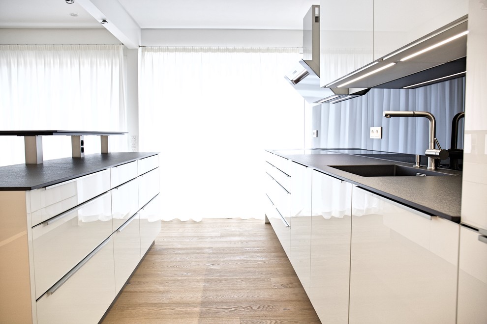 Photo of a large contemporary l-shaped open plan kitchen in Berlin with white cabinets, solid surface benchtops, black splashback, glass sheet splashback, with island, flat-panel cabinets, black appliances, light hardwood floors, beige floor and black benchtop.