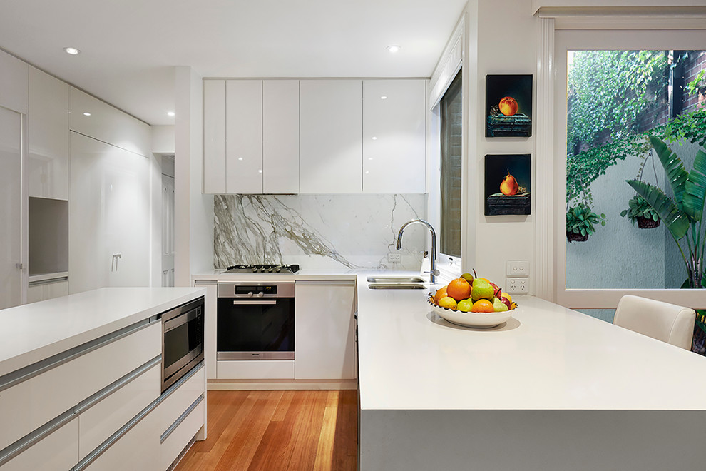 Photo of a mid-sized traditional l-shaped open plan kitchen in Melbourne with an undermount sink, flat-panel cabinets, white cabinets, marble benchtops, grey splashback, stone slab splashback, stainless steel appliances, light hardwood floors and multiple islands.