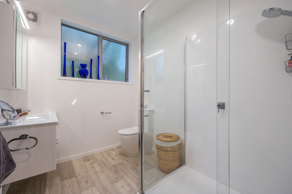 Photo of a small eclectic ensuite bathroom in Auckland with white cabinets, a corner shower, a one-piece toilet, white tiles, ceramic tiles, white walls, vinyl flooring, a wall-mounted sink, brown floors, a hinged door, white worktops, a single sink and a floating vanity unit.