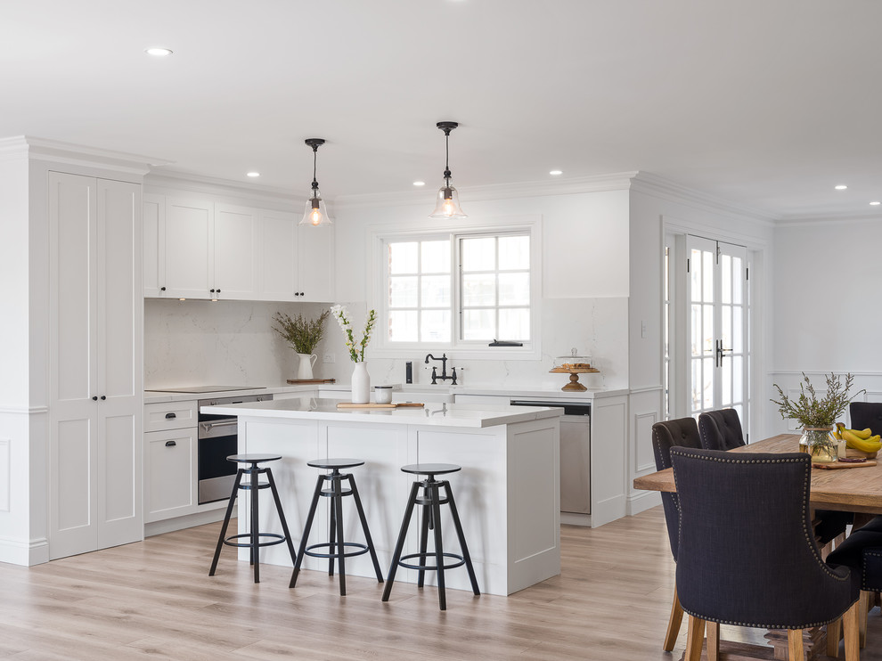 Mid-sized traditional l-shaped open plan kitchen in Sydney with a farmhouse sink, shaker cabinets, white cabinets, marble benchtops, white splashback, marble splashback, stainless steel appliances, laminate floors, with island, brown floor and white benchtop.